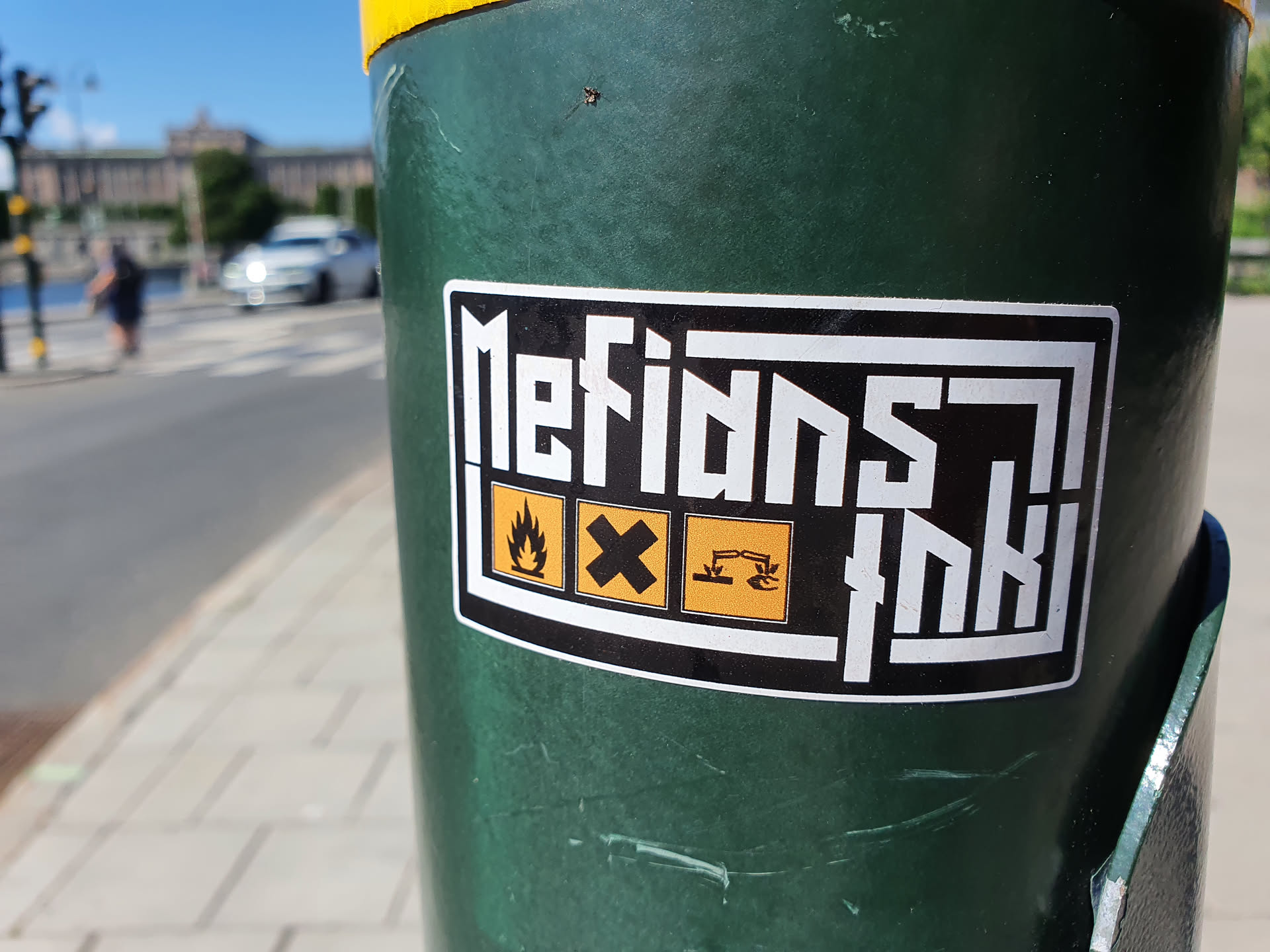 A black and white sticker with the text "Metians Ink" in a stylized font. It also features three small hazard symbols: fire, biohazard, and corrosive. The sticker is affixed to a green pole.