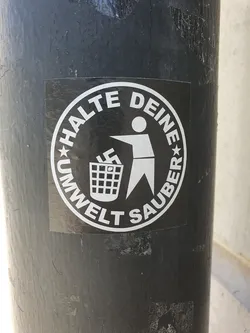 A black and white circular sticker with a figure throwing a swastika symbol into a trash can. The text around the circle says "Halte Deine Umwelt Sauber" which translates to "Keep your environment clean" in German.