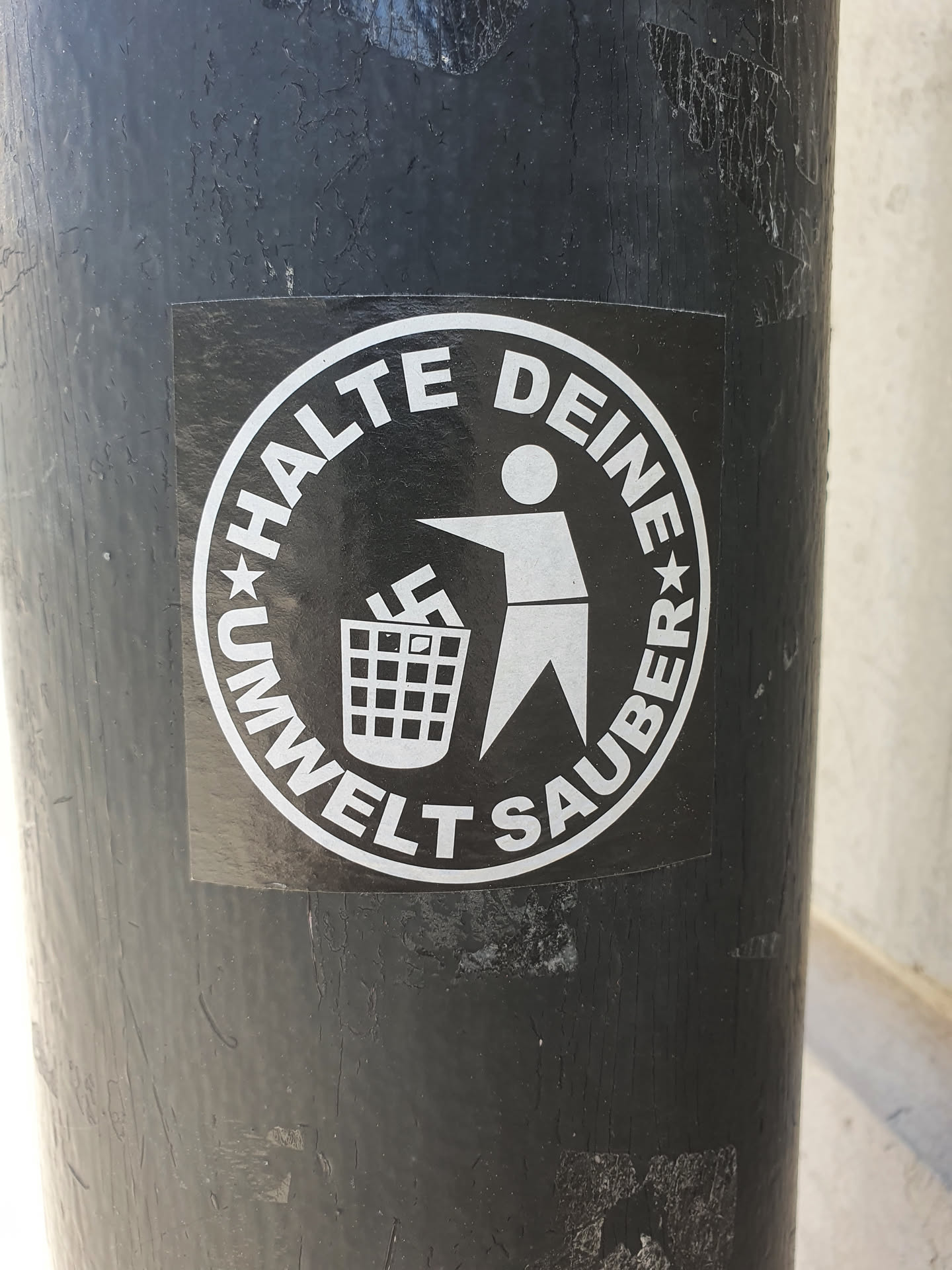 A black and white circular sticker with a figure throwing a swastika symbol into a trash can. The text around the circle says "Halte Deine Umwelt Sauber" which translates to "Keep your environment clean" in German.