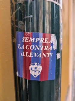 A weathered Levante UD sticker is affixed to a ribbed, dark green pole. The sticker features a bold, sans-serif typeface in white against a background of alternating vertical stripes in red and blue. The text reads "SEMPRE LA CONTRA ¡LLEVANT!" Beneath the text is the Levante UD crest. The sticker is partially peeling and shows signs of wear.