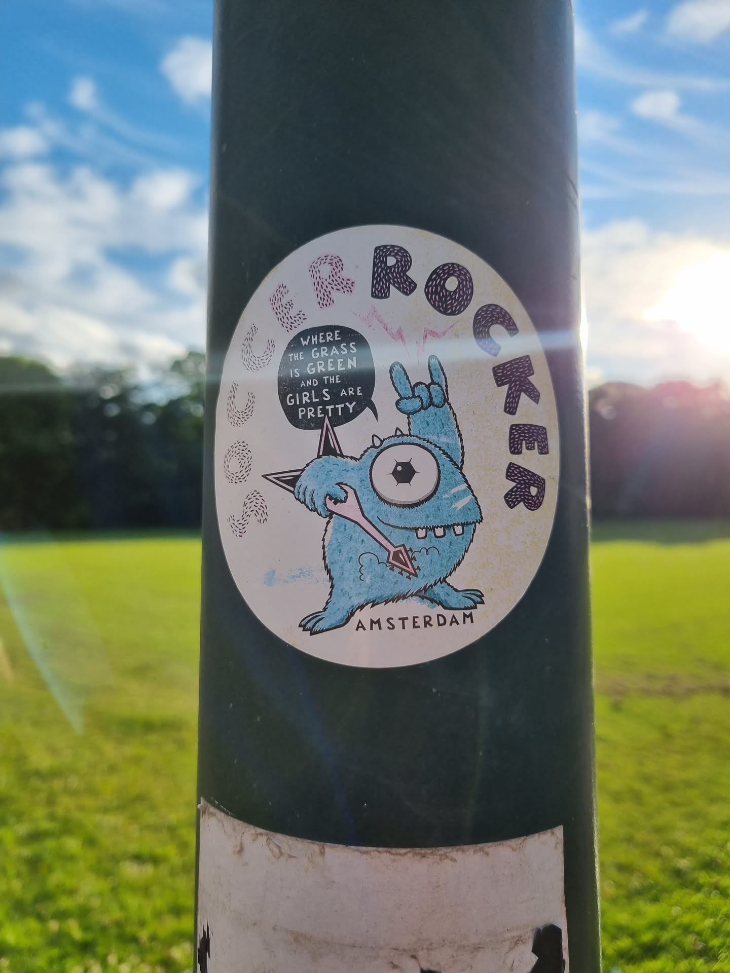 A round sticker featuring a cartoon monster playing a guitar. The text on the sticker says "Where the grass is green and the girls are pretty."  The sticker also has the word "Rocker" written in a circular pattern around the monster.  The sticker is affixed to a green pole.