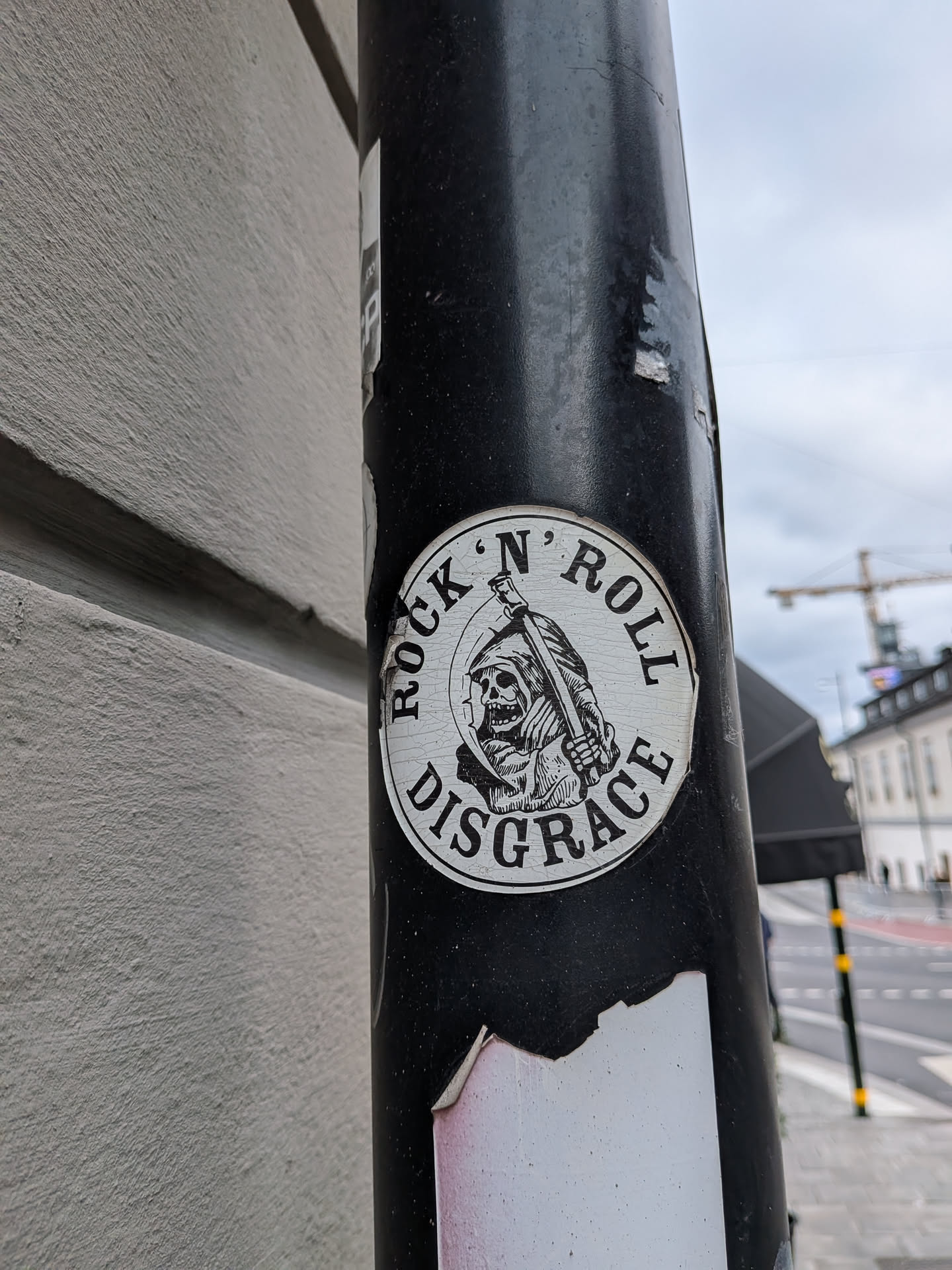 A circular sticker with a black border and a white interior. The sticker depicts a stylized Grim Reaper in a hooded cloak, holding a scythe over its shoulder. The Grim Reaper has a grinning skull face with visible teeth. The text 'ROCK' N' ROLL' is written in an arc above the Grim Reaper, and 'DISGRACE' is written in an arc below it. The artwork is rendered in a black and white, linework style, with a slightly distressed or cracked appearance to the white background.