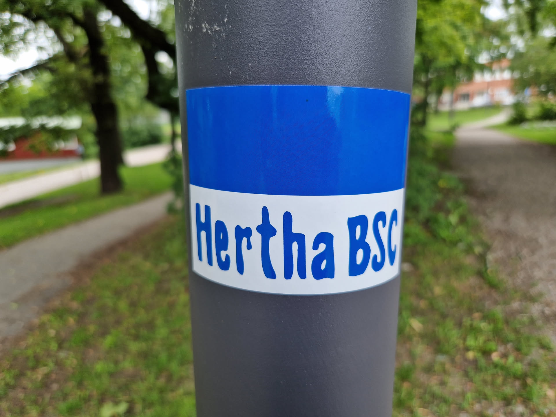A close-up shot of a blue and white Hertha BSC sticker affixed to a gray pole. The sticker features the club's name in a stylized font. The background is blurred, showing a park-like setting with trees and a path.