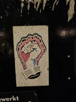 A sticker with a fist in the center and text that reads "Students for Justice in Palestine." The fist is depicted in pink and blue, with the text wrapped around it. There is an open book underneath the fist.