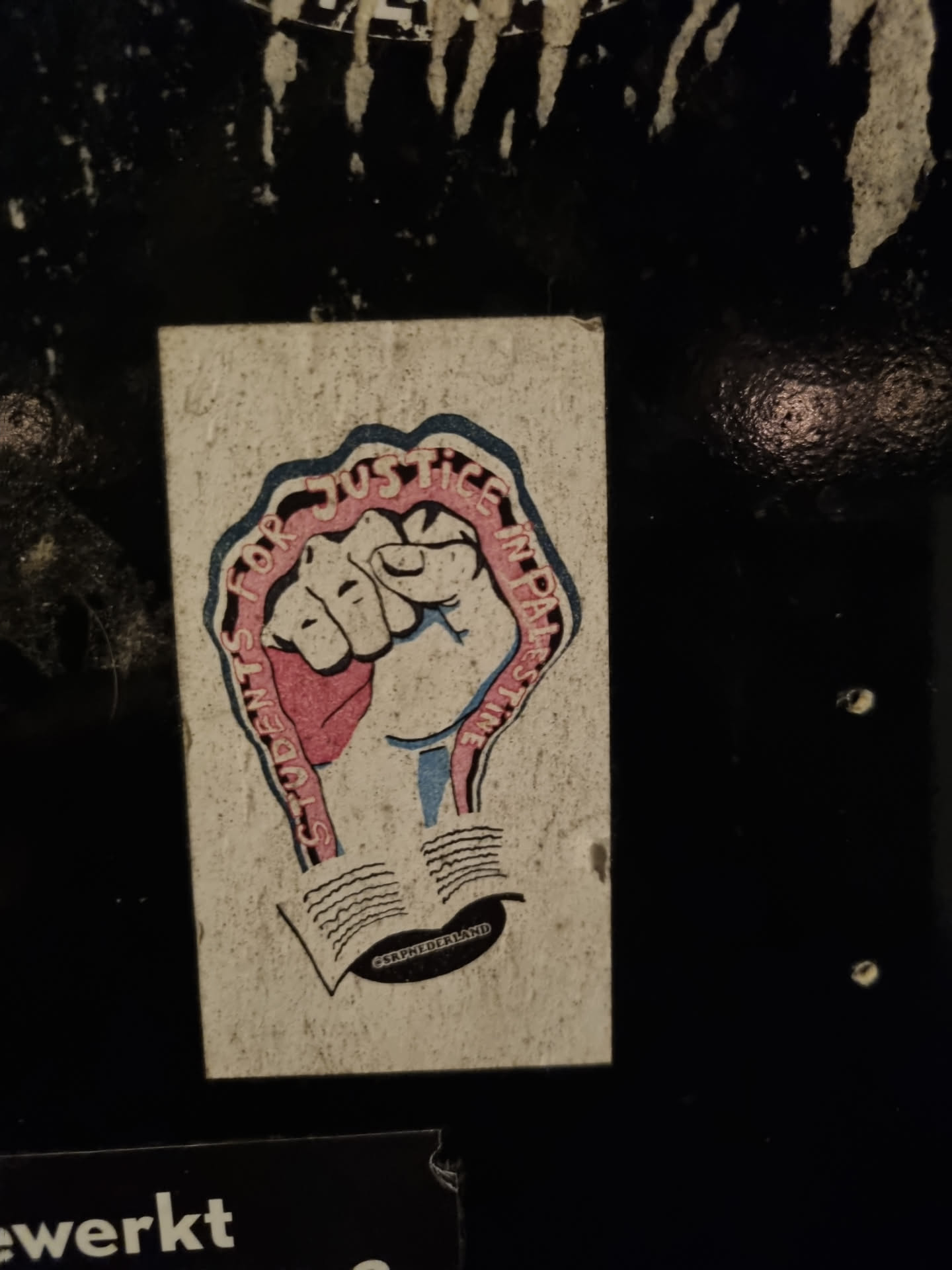 A sticker with a fist in the center and text that reads "Students for Justice in Palestine." The fist is depicted in pink and blue, with the text wrapped around it. There is an open book underneath the fist.