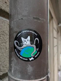 Round sticker featuring a cartoon cat sitting on the Earth, surrounded by an octopus's tentacles, set against a starry black background. The text 'NOTHING BEYOND REACH' is inscribed around the image, and 'LYDIA + STEFAN 2023-24' is written at the top. A small rover is depicted at the bottom.