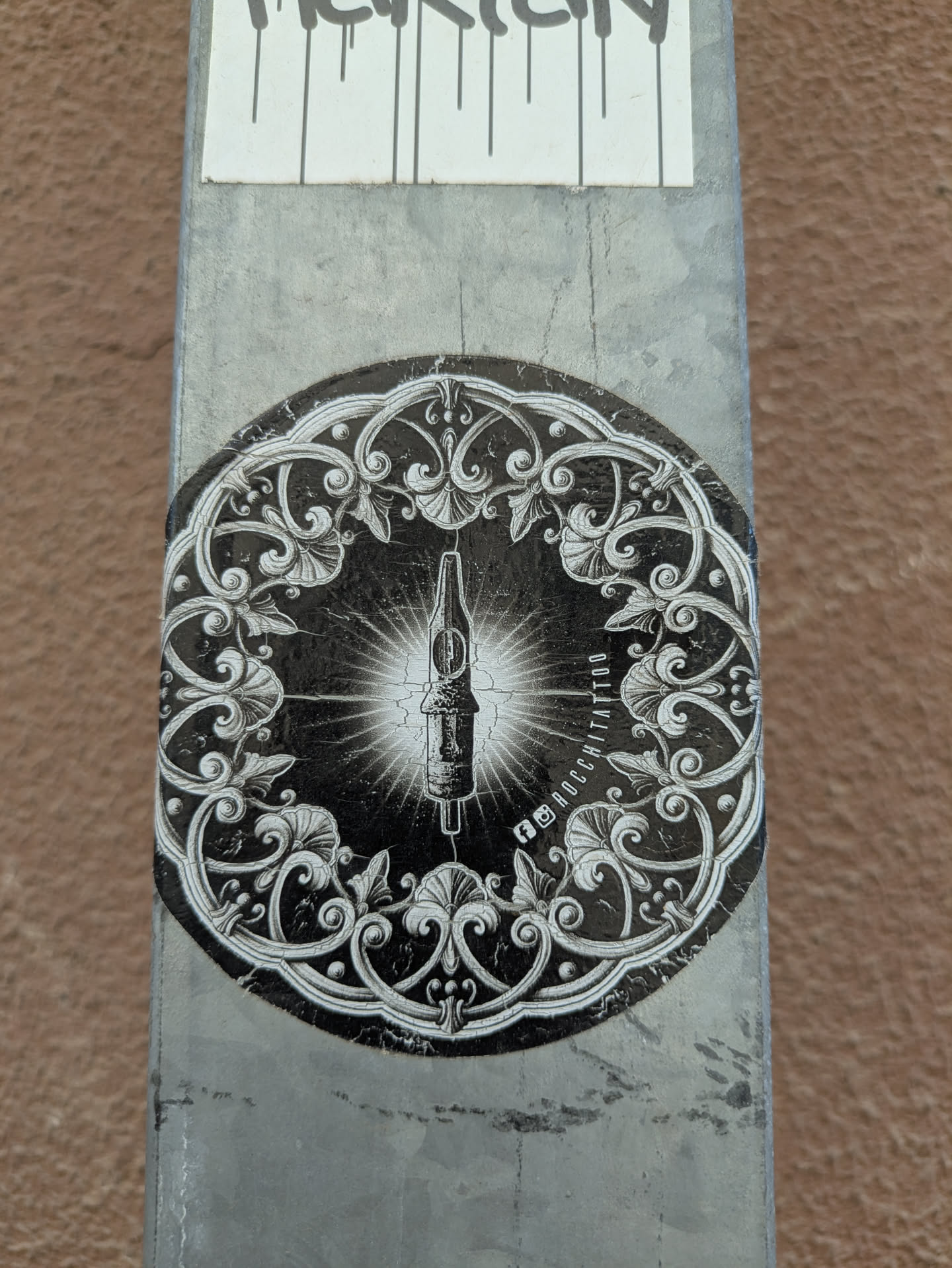 A round black and white sticker features a tattoo machine in the center, surrounded by an ornate, swirling floral design. Text around the right side indicates social media links.