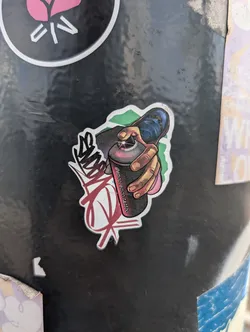 This sticker shows a hand holding a spray paint can. The can is dark grey with a pink stripe where the text "@enco_enco.tattoo" is written. The hand is pressing the spray nozzle. There is a stylized tag on the left side of the image, done in black and red. A green and light pink outline surrounds the sticker.