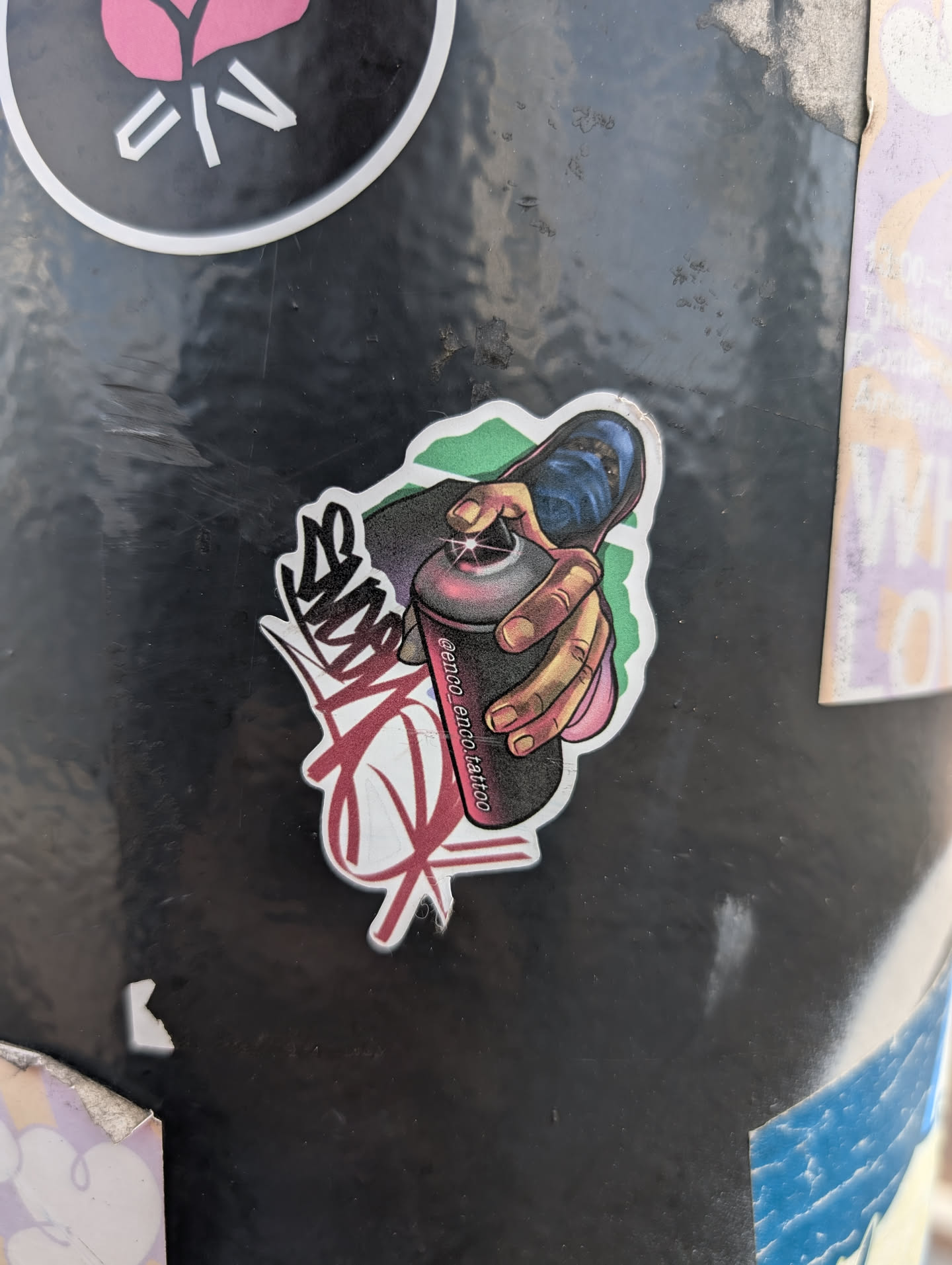 This sticker shows a hand holding a spray paint can. The can is dark grey with a pink stripe where the text "@enco_enco.tattoo" is written. The hand is pressing the spray nozzle. There is a stylized tag on the left side of the image, done in black and red. A green and light pink outline surrounds the sticker.