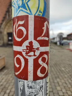 A red and white sticker with the numbers 6198 and a small devilish figure in the center. The sticker is affixed to a pole with other stickers around it.