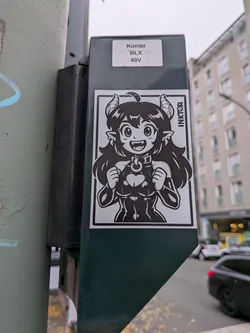 Street sticker A rectangular monochrome sticker showcasing a cheerful, anime-style demon or succubus character. She features large expressive eyes, fangs visible in an open smile, pointed ears, and two curved horns emerging from thick dark hair. She wears a collar with a ring and a form-fitting outfit featuring a heart-shaped chest cutout, posing with clenched fists raised in an excited gesture. Vertically along the upper-right inner border, the artist's name 'INKTOR' is displayed.