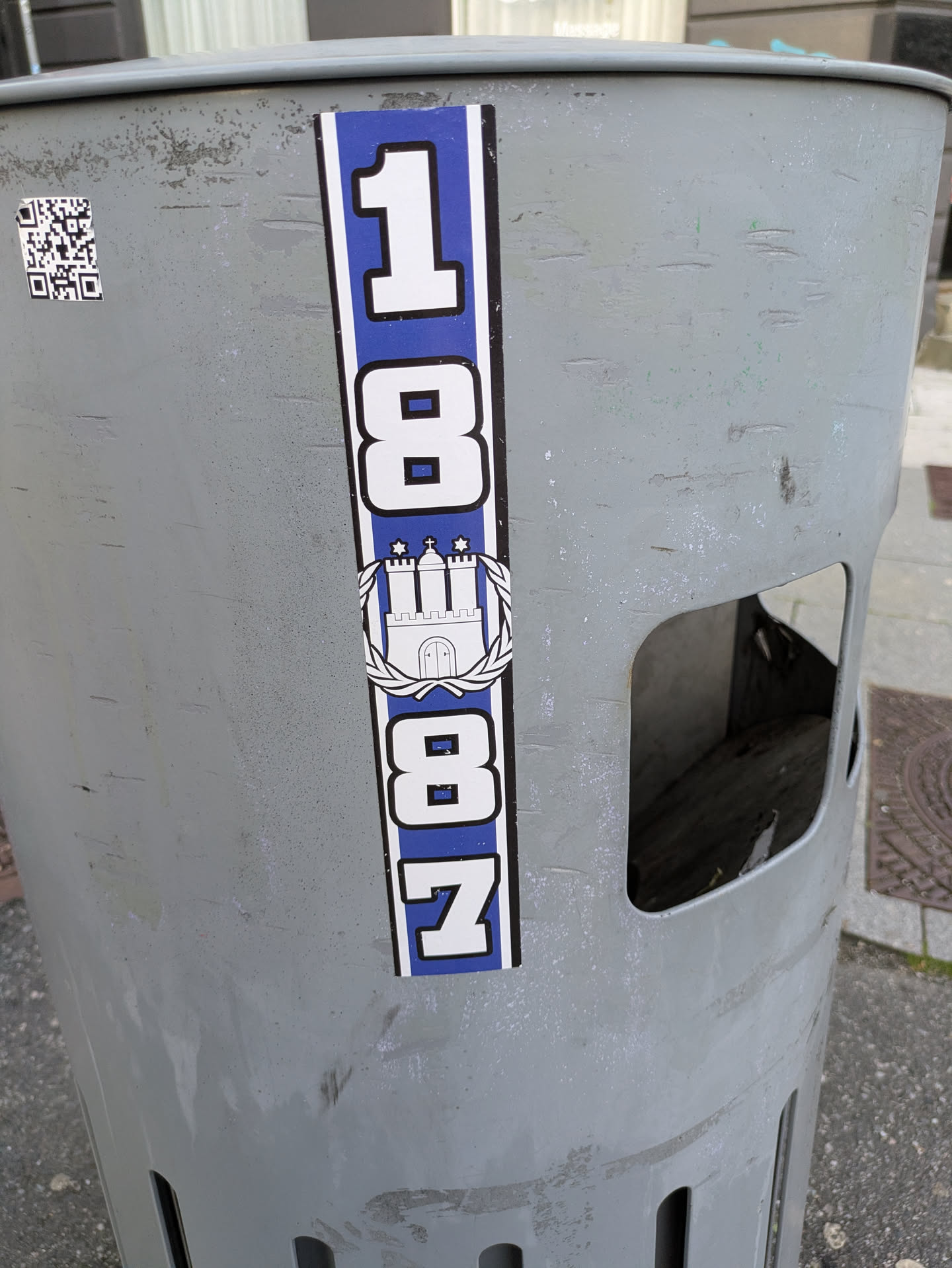 The central sticker is vertical, predominantly blue and white. It displays the numbers '18' at the top, followed by a white crest of a castle with three towers (the Hamburg city crest) encircled by a wreath. Below the crest are the numbers '87'. The entire design is set against a blue background with a white border. The sticker is placed on a worn, light grey metal trash bin that has visible scratches and marks. To the left of the main sticker, a somewhat damaged QR code is visible.
