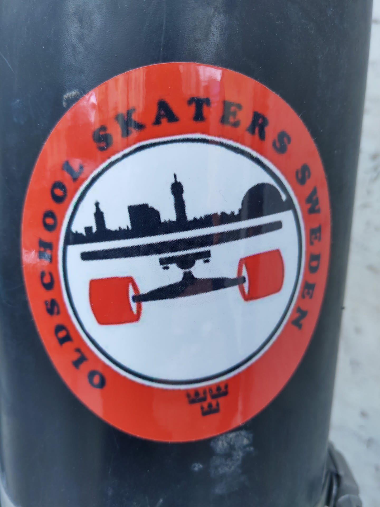 A round sticker with an orange border and a white center. The text "Oldschool Skaters Sweden" is written around the edge. In the center is a black silhouette of a skateboard with a city skyline in the background. There is a small logo at the bottom.