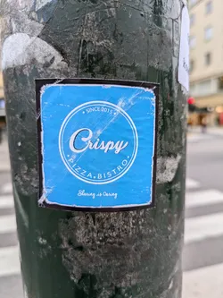 A blue sticker on a pole advertising Crispy Pizza Bistro, established since 2015. The sticker has a simple design with the restaurant's name and slogan.