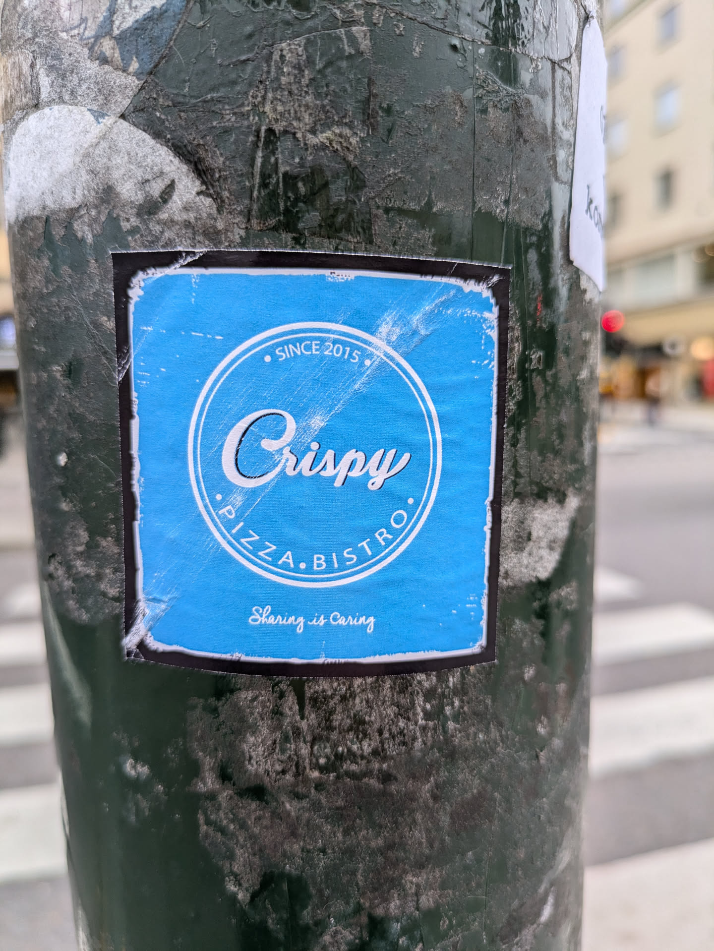 A blue sticker on a pole advertising Crispy Pizza Bistro, established since 2015. The sticker has a simple design with the restaurant's name and slogan.