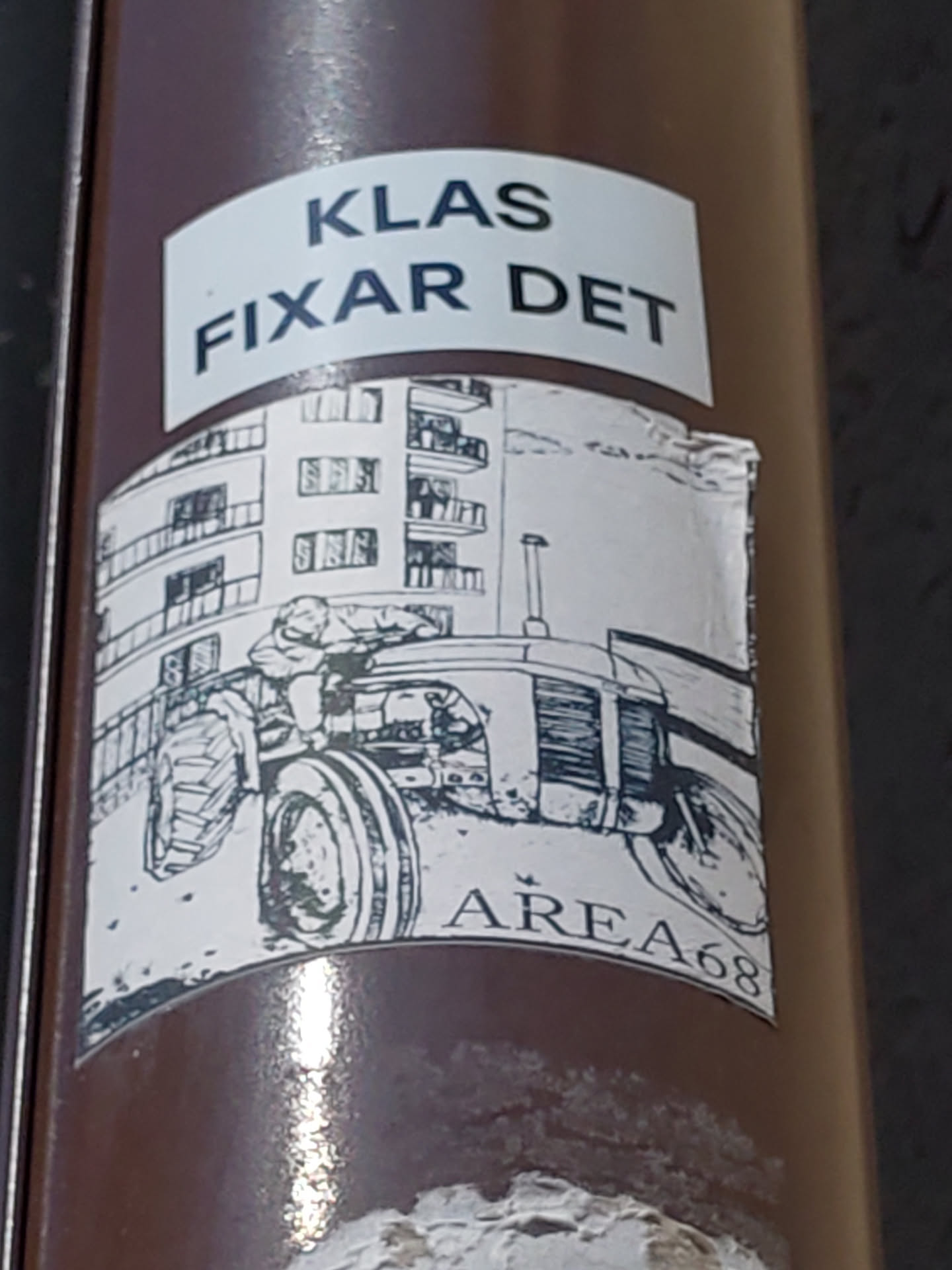 A sticker featuring a black and white drawing of a tractor in front of an apartment building. The text 'KLAS FIXAR DET' is visible at the top, and 'AREA68' is at the bottom.