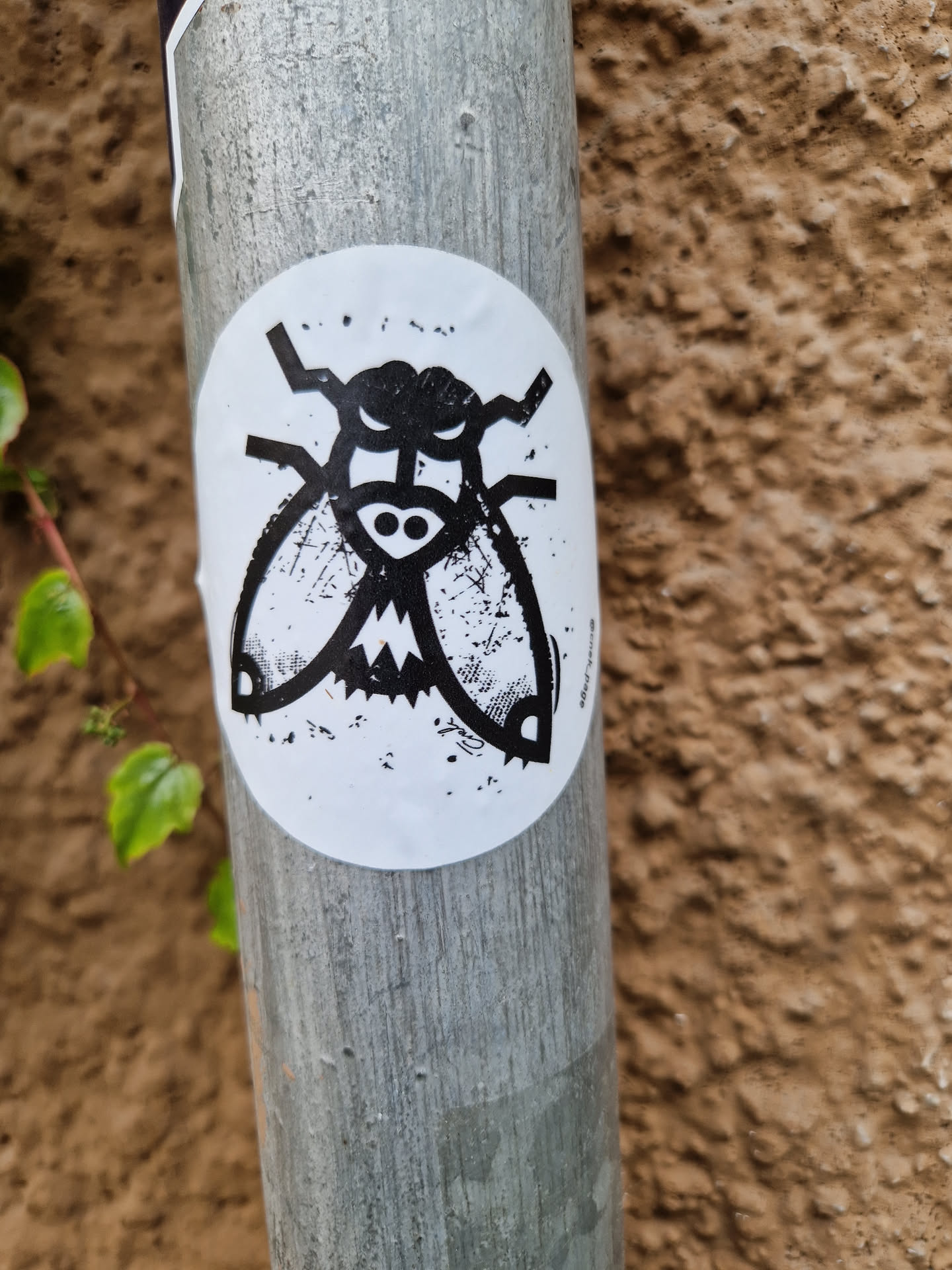 A round sticker with a black and white illustration of a fly with angry expression. It is attached to a gray metal pole against a brown textured background. The sticker has a slightly distressed or grunge look.