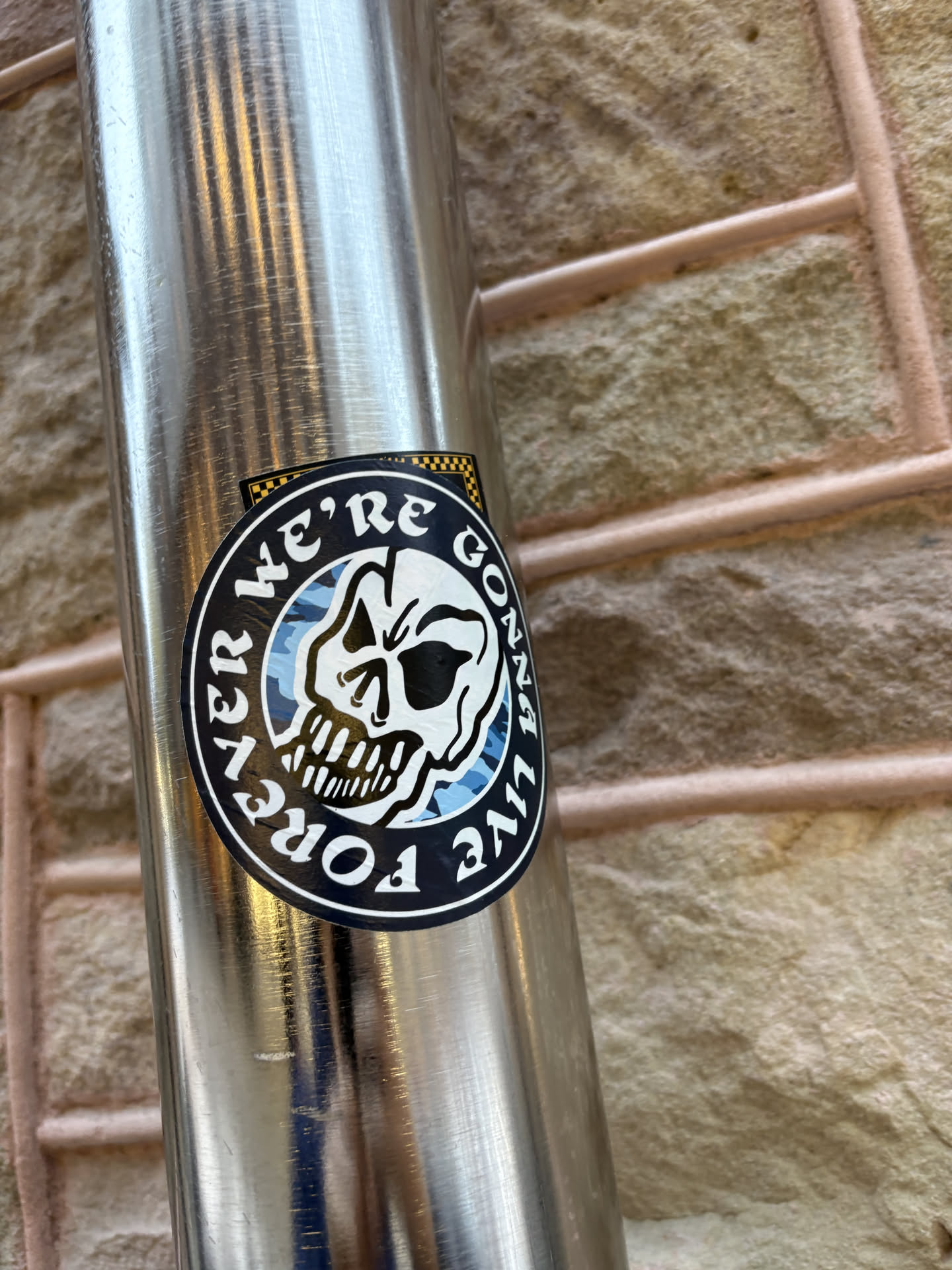 A circular sticker featuring a stylized white skull with black outlines and shadows, set against a blue and light blue camouflage pattern. The skull is centered within a thick black border that contains white block letters spelling "FOREVER WE'RE GONNA LIVE" in an arc. A small black and yellow checkered pattern is visible at the very top edge, partially obscured by the main circular design.
