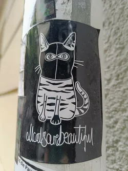 Black and white sticker featuring a stylized illustration of a cat wearing what looks like a mask or balaclava. The cat is depicted sitting down, and the sticker has the text "all cats are beautiful" written at the bottom.