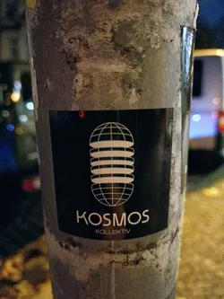 Black and white sticker with a globe-like design and the text Kosmos Kollektiv. The sticker is attached to a weathered gray pole.