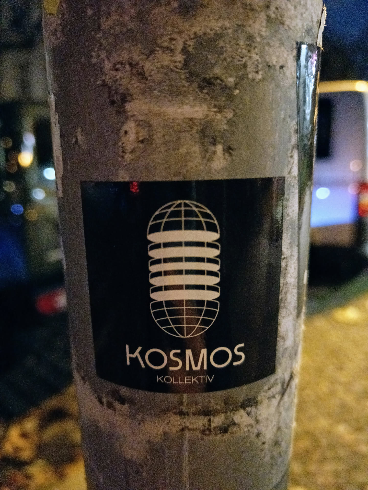 Black and white sticker with a globe-like design and the text Kosmos Kollektiv. The sticker is attached to a weathered gray pole.