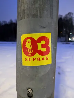 A rectangular sticker with a bright yellow background, affixed to a grey metal pole. The primary design elements are in a bold red color. On the left side, there is a stylized profile of a person's head, facing right, with an open mouth as if singing or shouting. The figure appears to be wearing a striped scarf around their neck. To the right of the head, the large numbers '03' are prominently displayed. Below the '03' and partially below the head, the word 'SUPRAS' is written in capital letters. The sticker shows minor wear, including a slight wrinkle and a small tear on its top left corner.