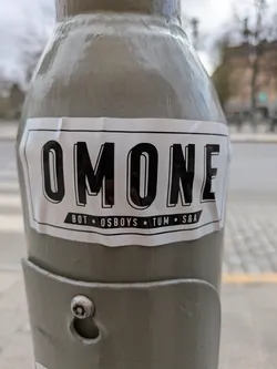 This is a white rectangular sticker on a light post. The sticker is printed with the text "OMONE" in bold black letters at the top. Underneath that are the words "BOT • OSBOYS • TUM • S&A" in smaller black letters.