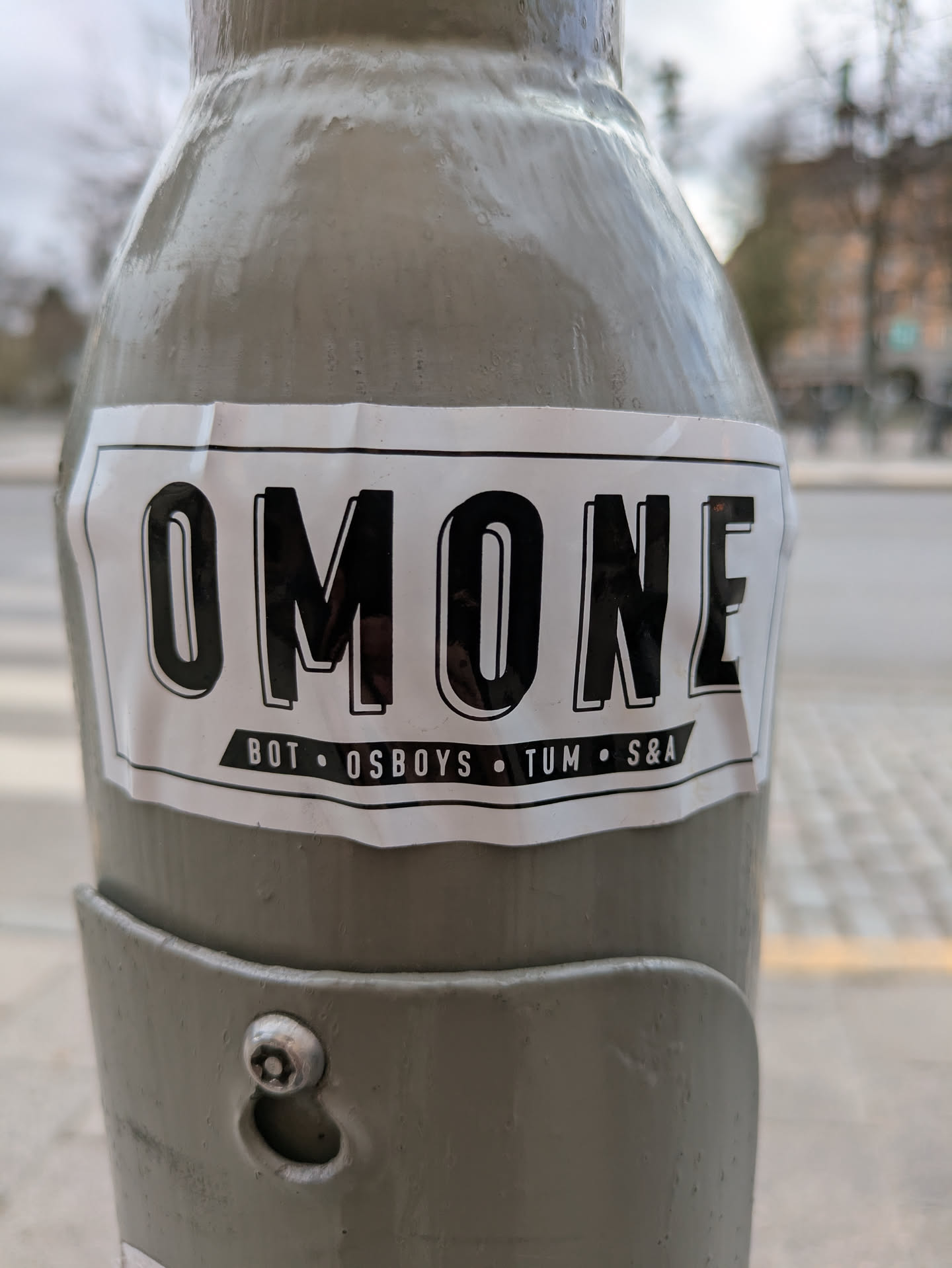 This is a white rectangular sticker on a light post. The sticker is printed with the text "OMONE" in bold black letters at the top. Underneath that are the words "BOT • OSBOYS • TUM • S&A" in smaller black letters.