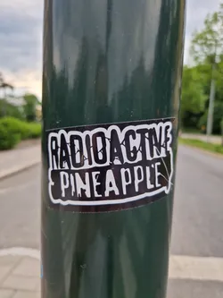 A sticker featuring the words "Radioactive Pineapple" in a stylized font. The sticker is black and white and appears to be affixed to a dark green pole.