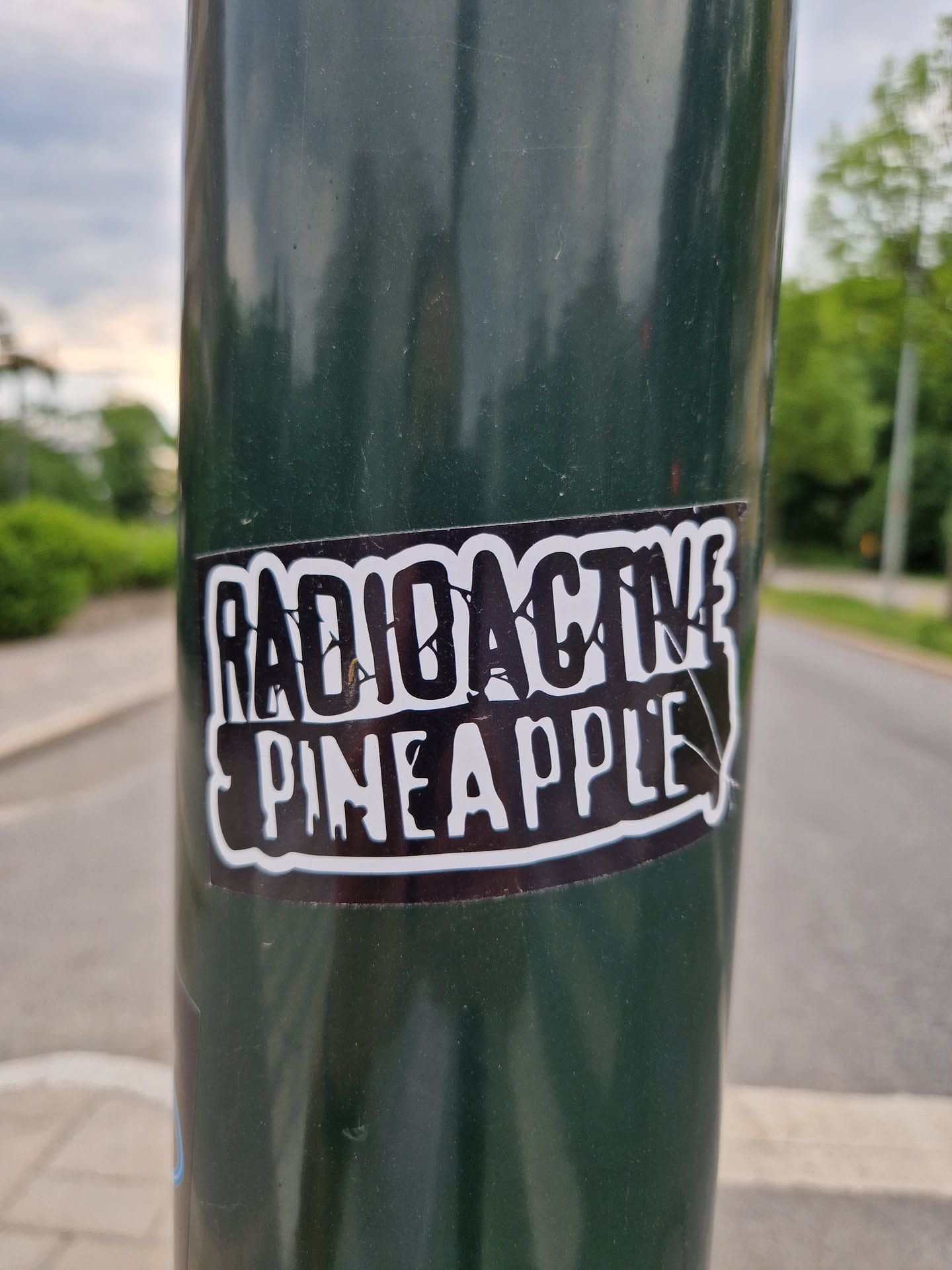 A sticker featuring the words "Radioactive Pineapple" in a stylized font. The sticker is black and white and appears to be affixed to a dark green pole.