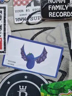 A sticker featuring an illustration of a purple eggplant with bat-like wings. The sticker has a blue border and text in the bottom-left corner that seems to be social media handles. 