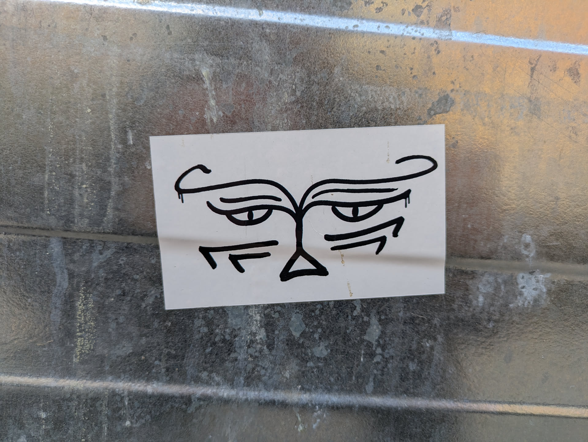 A black and white sticker depicting a minimalist, cartoonish face with a distinctly grumpy or annoyed expression. The face features arched eyebrows, narrow eyes, and a triangular nose. The overall style is reminiscent of quick sketches or doodles.
