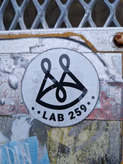 A round, white sticker with a black logo. The logo appears to be an eye with stylized lines above it. The text ". LAB 259." is visible below the logo.