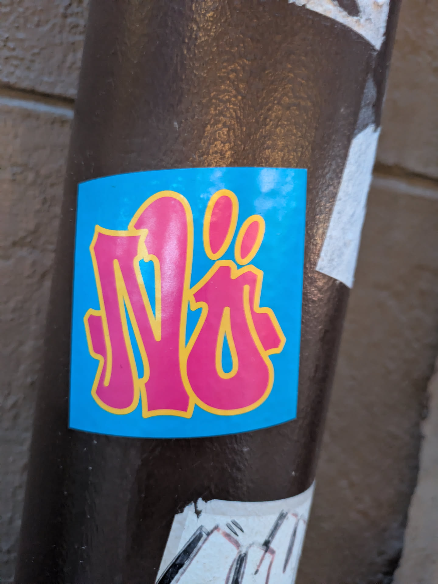 The sticker is predominantly a bright, sky blue. Centered on this blue background are stylized, bubble-like letters rendered in a vibrant pink outline with a yellow inner border. The letters appear to form a word or tag, with a distinct shape resembling an 'N' or a stylized monogram, followed by two small, oval shapes that could be dots or part of the lettering. The edges of the sticker are clean and appear to be slightly rounded.