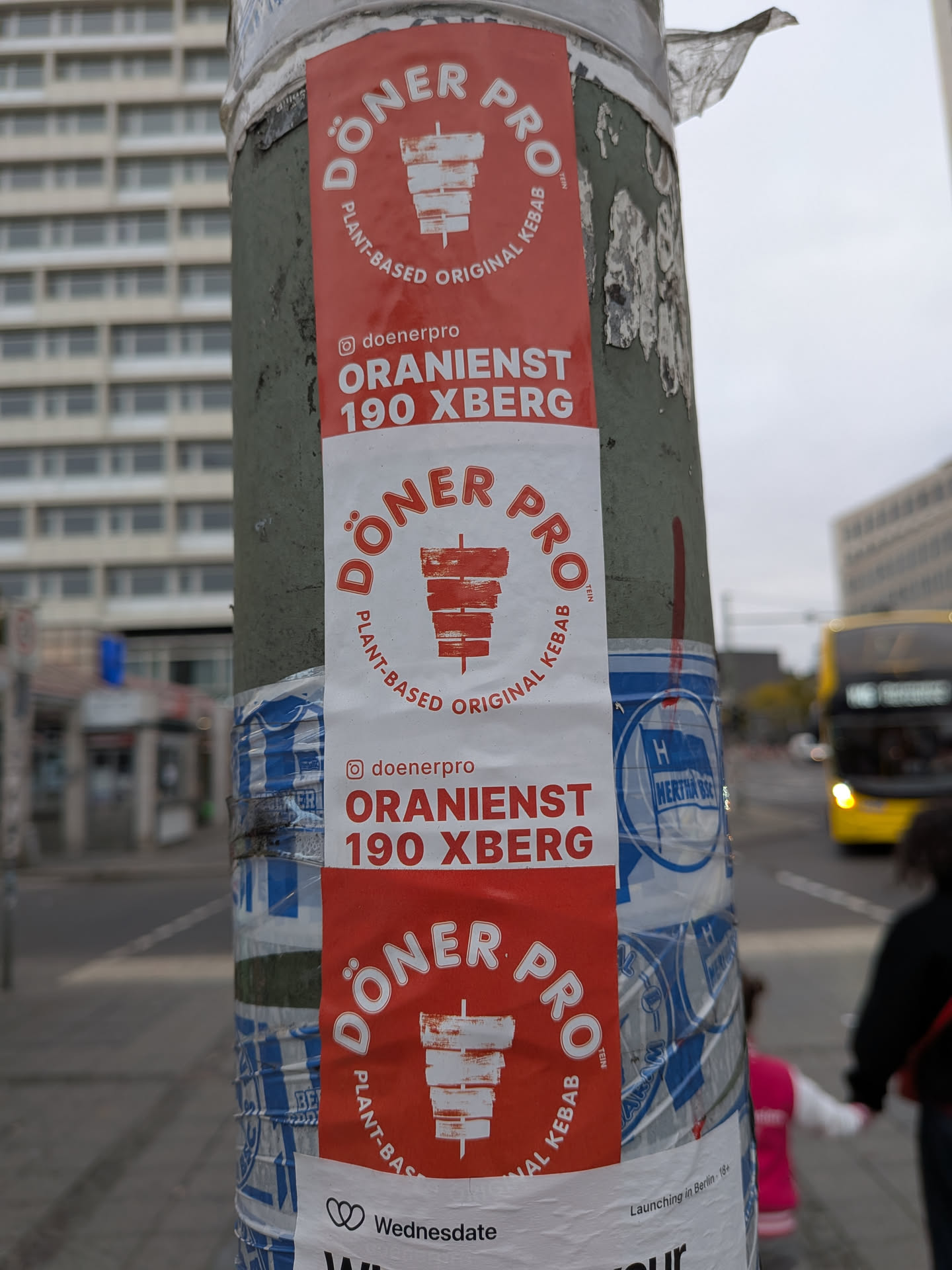 A vertical adhesive tape-style sticker strip affixed to a weathered metal street pole. The design consists of alternating red-on-white and white-on-red repeating blocks promoting 'DÖNER PRO'. Each section prominently displays the circular logo featuring a döner kebab skewer graphic in the center, encircled by 'DÖNER PRO' at the top and 'PLANT-BASED ORIGINAL KEBAB' underneath. Below the circular mark, an Instagram icon accompanies the handle '@doenerpro' alongside the street location 'ORANIENST 190 XBERG'.