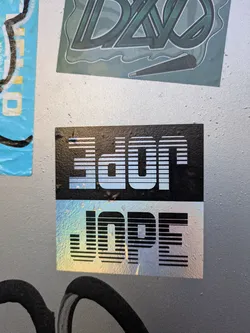 A holographic sticker with the word "DOPE" stacked on top of "JOPE" in a retro style font. The sticker has a black background and a iridescent effect.