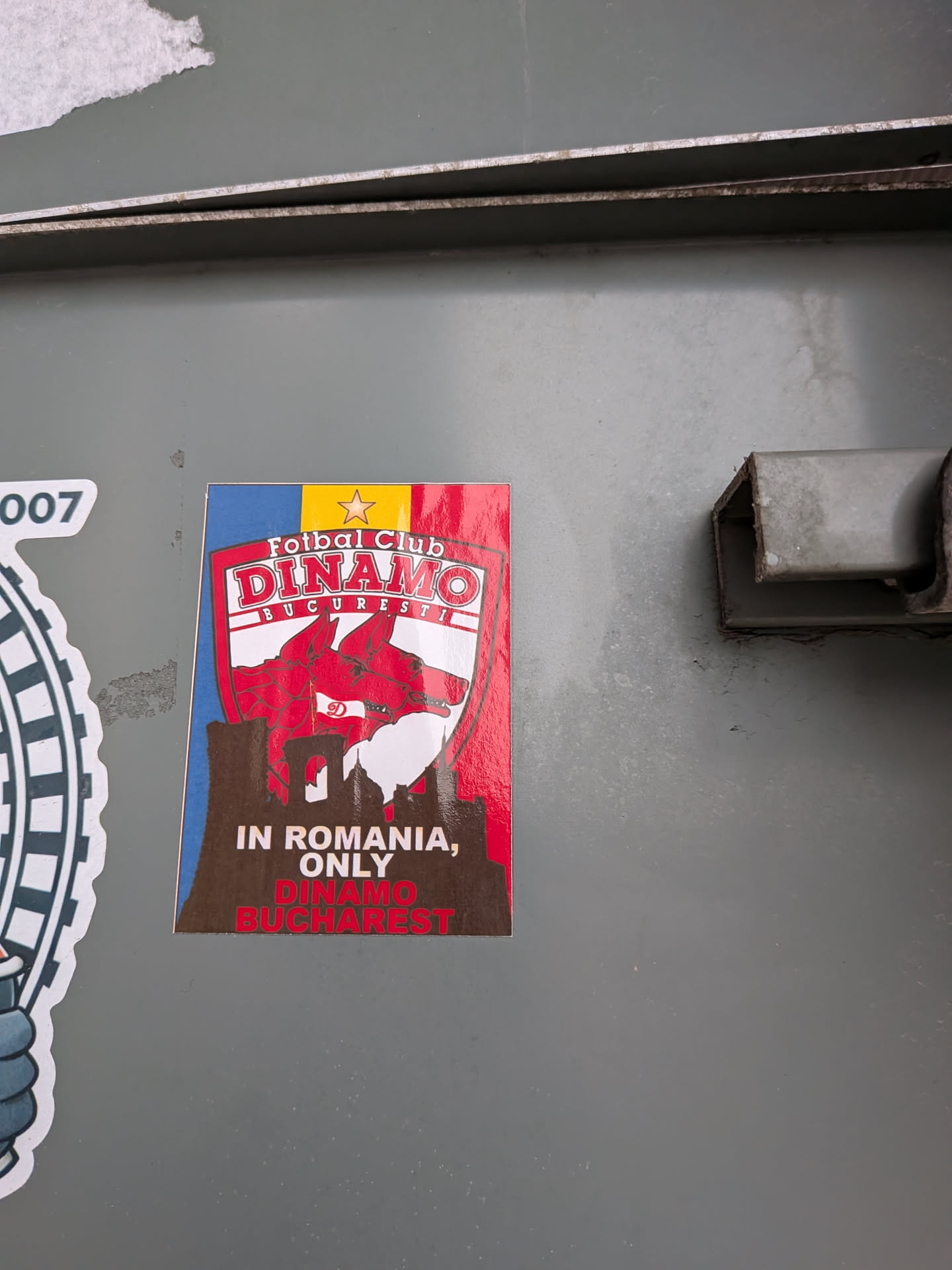 A vertical rectangular sticker featuring the crest of Romanian football club 'Fotbal Club Dinamo București', adorned with red dog heads and the club emblem, set against the tricolor backdrop of the Romanian flag (blue, yellow with a gold star, and red). Below the crest, a brown silhouette of Bucharest city landmarks is displayed with the bold text 'IN ROMANIA, ONLY DINAMO BUCHAREST' printed across it.