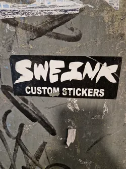 A black and white sticker with the word "SWEZINX" in a stylized font, and the words "CUSTOM STICKERS" underneath. The sticker is placed on a gray wall with various markings.
