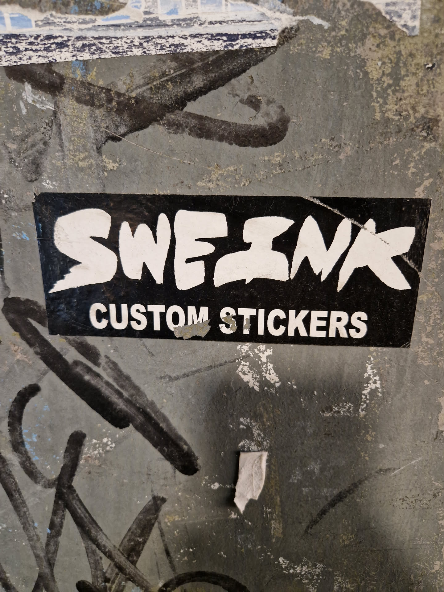 A black and white sticker with the word "SWEZINX" in a stylized font, and the words "CUSTOM STICKERS" underneath. The sticker is placed on a gray wall with various markings.