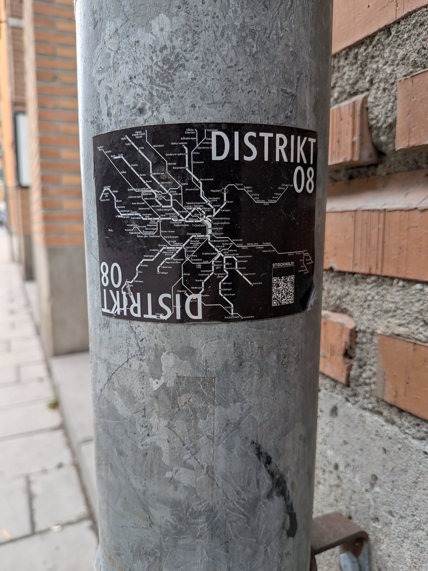 A rectangular black sticker adhered to a metal pole, featuring a white, stylized map of the Stockholm metro system. The map displays various lines and station names in white text against the black background. Prominently displayed in the upper right corner is the text 'DISTRIKT 08' in a bold, sans-serif white font. The same text, 'DISTRIKT 08', is repeated in the lower left corner, inverted and slightly obscured by wear. Above a small QR code in the bottom right, the word 'STOCKHOLM' is visible. The sticker shows minor signs of wear and tear, including slight peeling and scuffs.