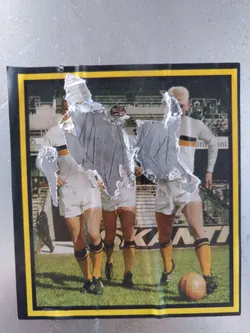 A damaged sticker featuring a retro photo of three football players in action. The sticker has a yellow border and shows some wear and tear.  The players are wearing white shorts and yellow and black striped socks.