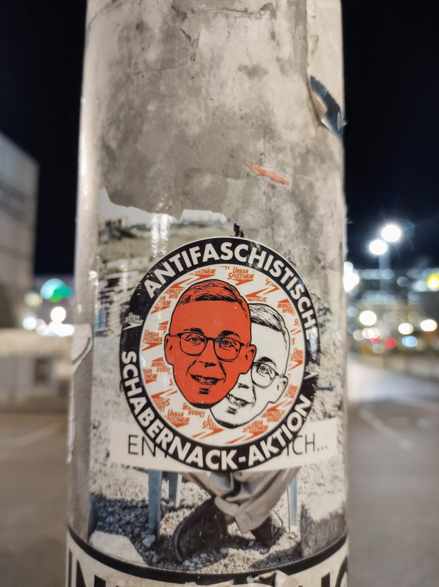 A round sticker with an anti-fascist theme. It features two faces, one orange and one white, possibly representing different perspectives or individuals. The text on the sticker reads "Antifaschistisch Schabernack-Aktion..." which translates to "Anti-fascist prank action...". The sticker has a slightly distressed look, indicating it might be placed outdoors.