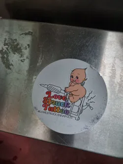 Round sticker featuring a cherubic baby riding a tattoo machine. The text "Love Struck Tattoo" is written in a rainbow-colored font.  The Instagram handle @lovestruckannapolis is also included.