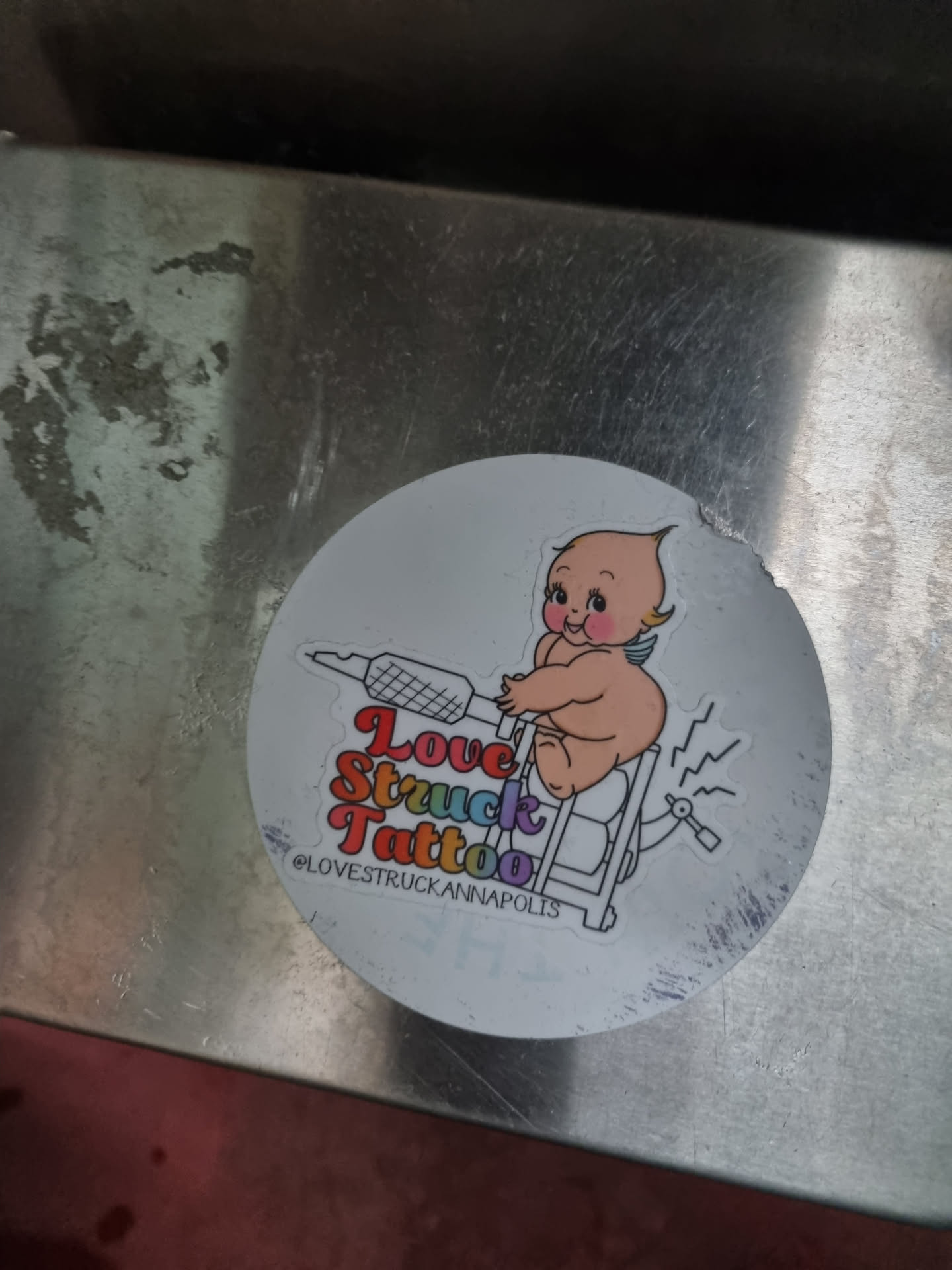 Round sticker featuring a cherubic baby riding a tattoo machine. The text "Love Struck Tattoo" is written in a rainbow-colored font.  The Instagram handle @lovestruckannapolis is also included.