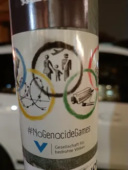 Sticker expressing opposition to the Olympic Games, possibly due to human rights concerns. It features a modified Olympic logo with barbed wire and surveillance imagery. The text includes the hashtag #NoGenocideGames and the logo for Gesellschaft für bedrohte Völker (Society for Threatened Peoples).