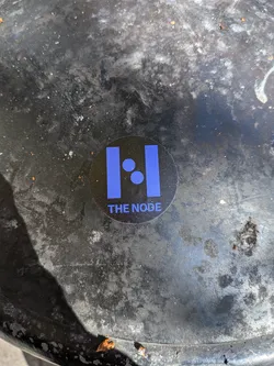 Street sticker A round black sticker affixed to a worn, textured dark surface. The sticker displays a bright blue graphic emblem consisting of two thick vertical rectangular bars with two diagonally aligned blue circles situated between them. Below this geometric icon, the text 'THE NODE' is printed in a clean, uppercase sans-serif typeface in the same blue color.