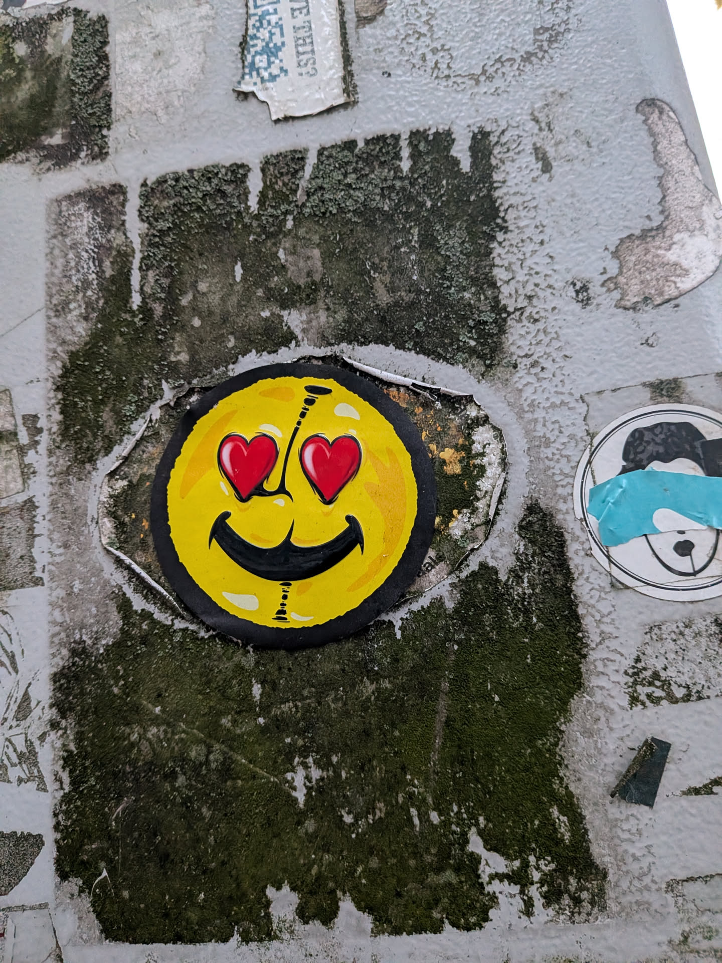 A circular sticker featuring an altered yellow smiley face. The emoji has bright red heart-shaped eyes with white light reflections, a wide black smiling mouth, and subtle black seam or stitch-like markings running down the center. The sticker is bordered in black and affixed over a weathered, mossy surface with peeling paper edges behind it.