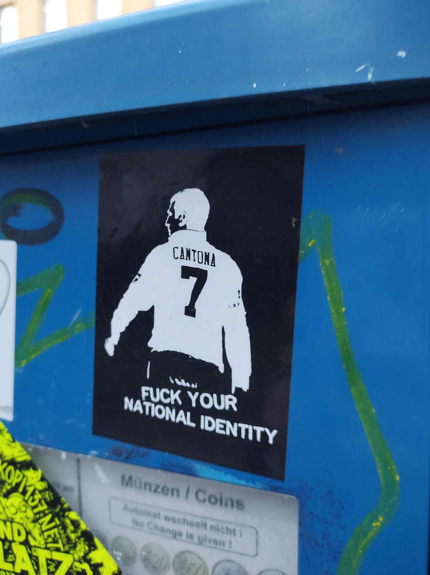 A sticker featuring a stylized image of Eric Cantona, the famous Manchester United footballer, with his name and number 7 on the back of his shirt. The text 'Fuck Your National Identity' is printed below the image. The sticker is affixed to a blue metal surface.