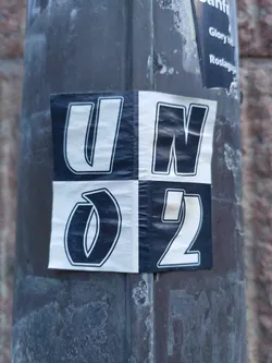 A sticker with a bold design featuring the letters UN and the numbers 02 arranged in a visually striking pattern. The sticker is affixed to a weathered gray pole.