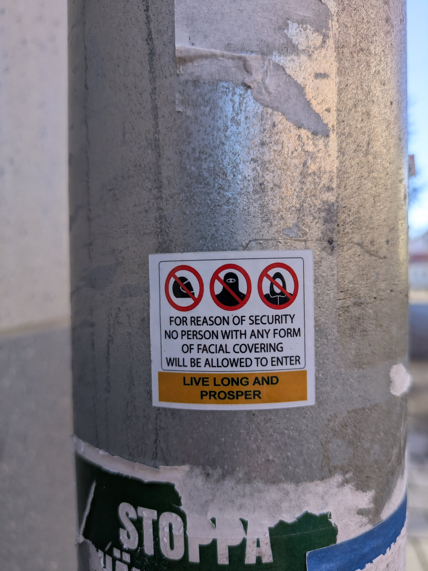 The sticker on the pole features three images with red circles and a line through them, depicting various forms of facial coverings that are not allowed. The text reads: "FOR REASON OF SECURITY NO PERSON WITH ANY FORM OF FACIAL COVERING WILL BE ALLOWED TO ENTER LIVE LONG AND PROSPER".